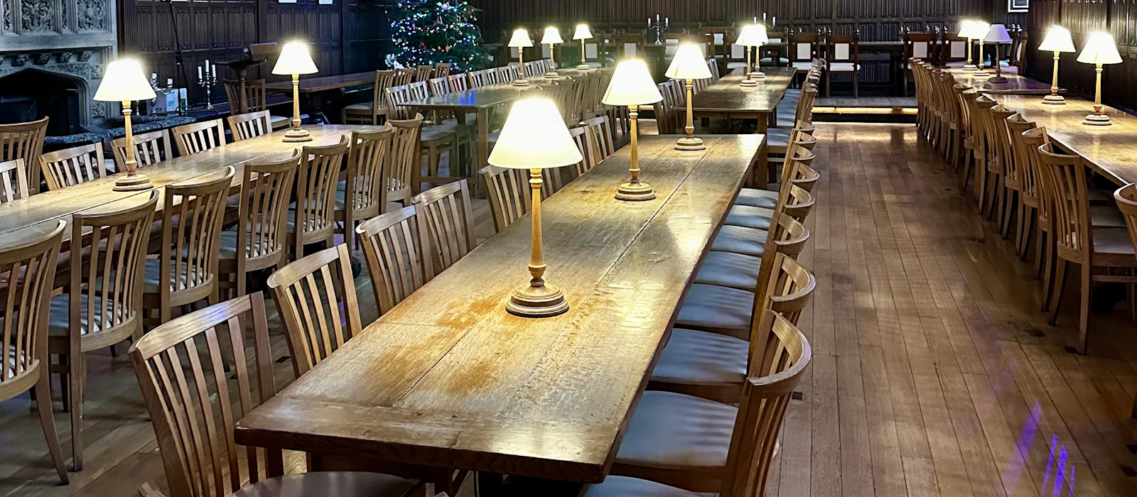 University dining hall with long wooden tables, table lamps, and high-backed chairs.