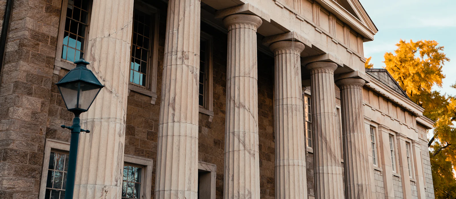 Stone government building with tall columns and a lamp post beside autumn trees.