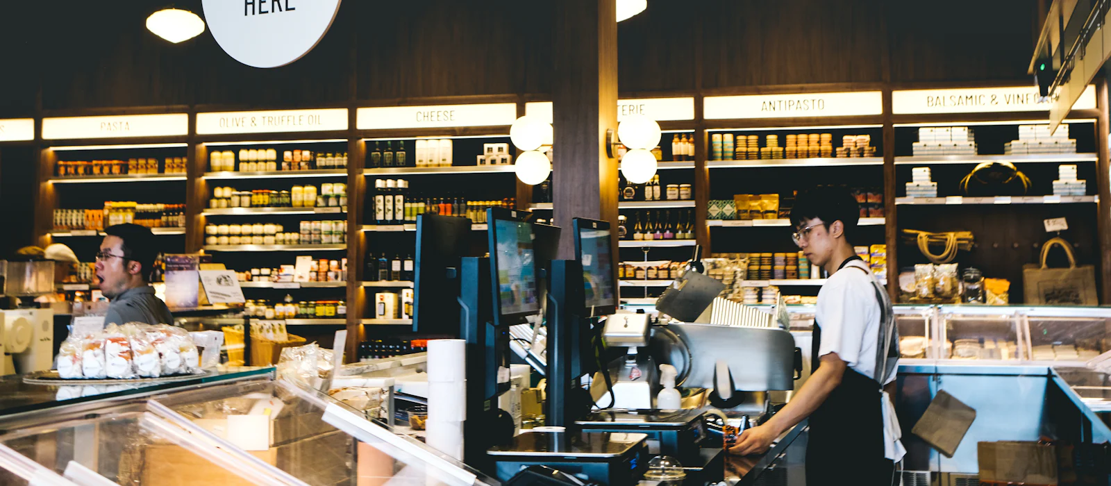 Grocery store cheese counter with shelves of specialty goods and staff at the registers.