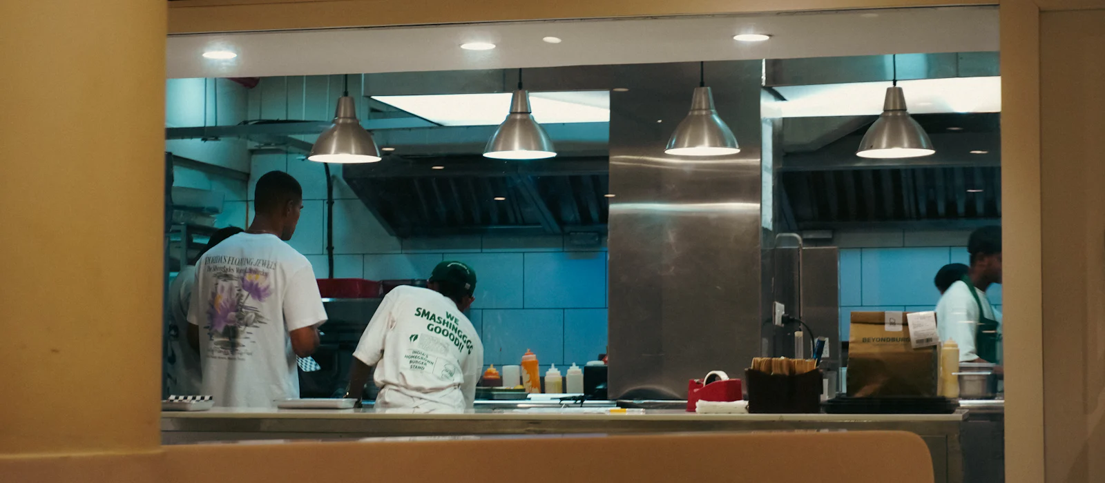 Two cooks working behind a kitchen pass-through window under pendant lights.