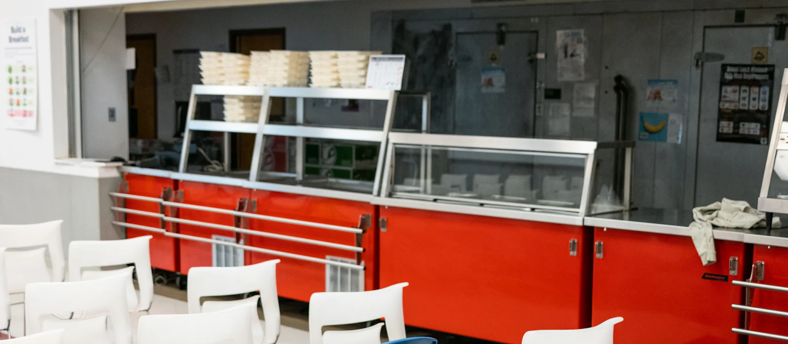 School cafeteria serving line with red counters and white chairs in front.