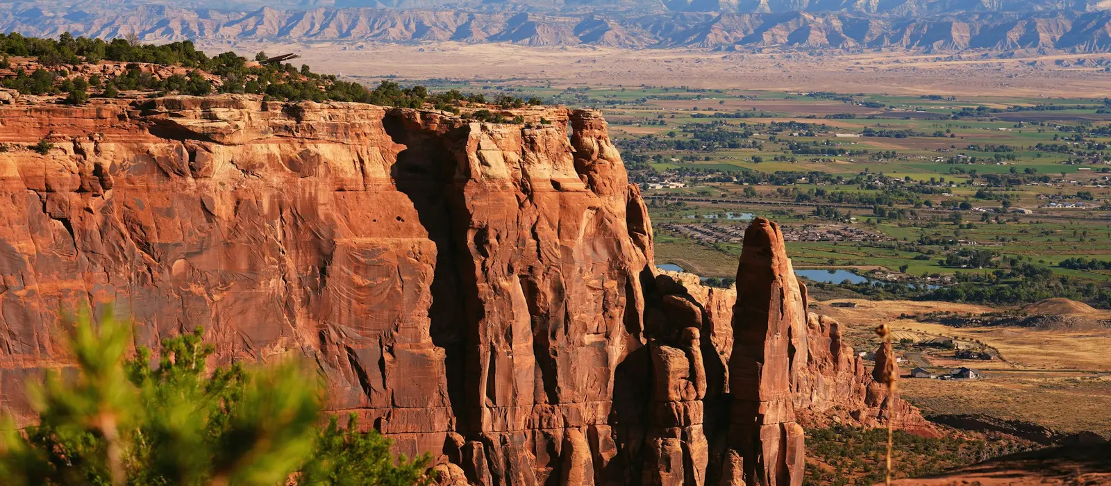 Red sandstone cliffs of Colorado National Monument above the Grand Valley farmland near Grand Junction, with the Book Cliffs on the horizon.