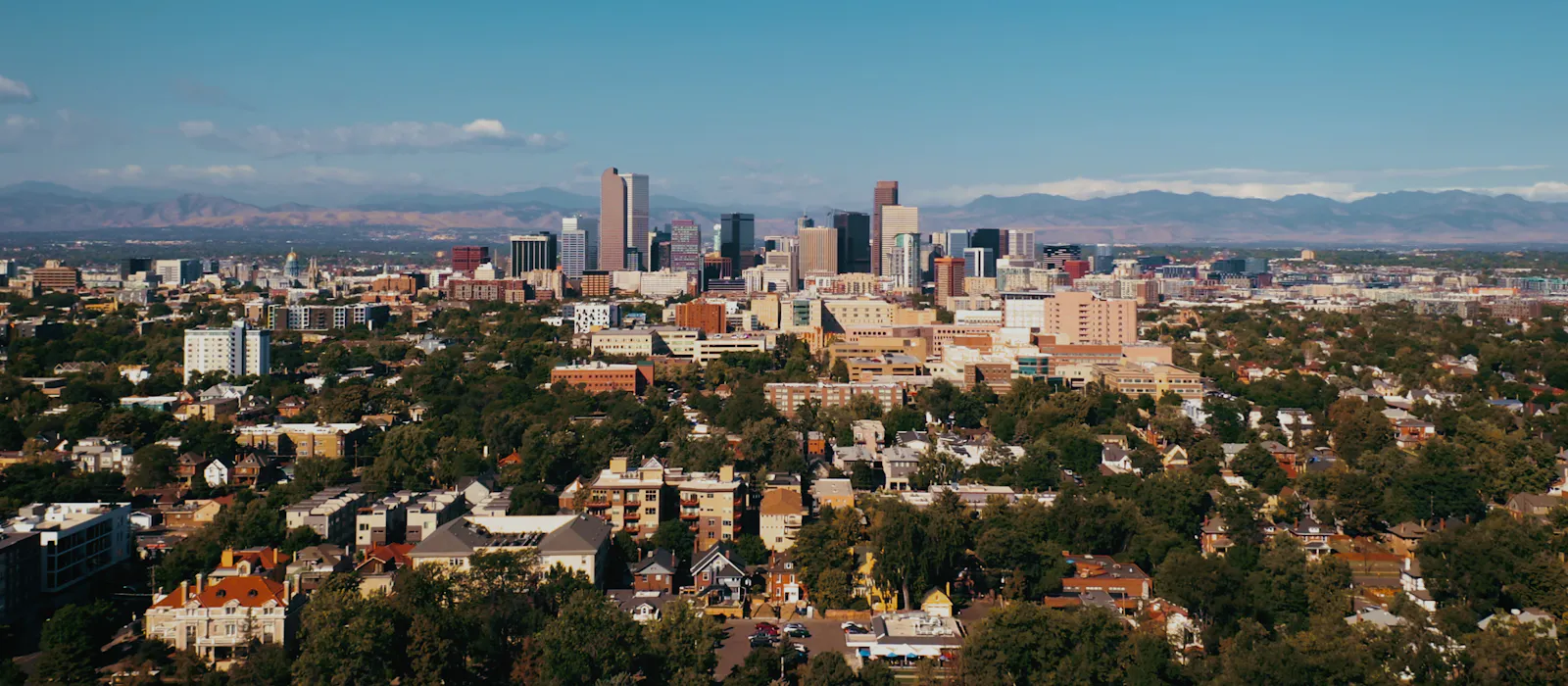 Downtown Denver skyline rising above tree-lined neighborhoods, with the Front Range on the horizon.