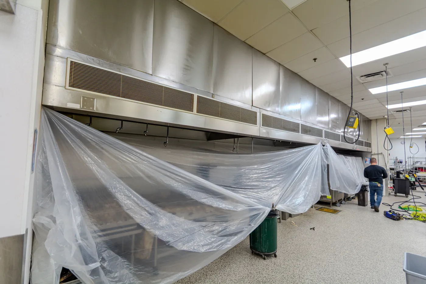 A long institutional kitchen hood with containment sheeting hung along its full length, funnelling into a collection drum.