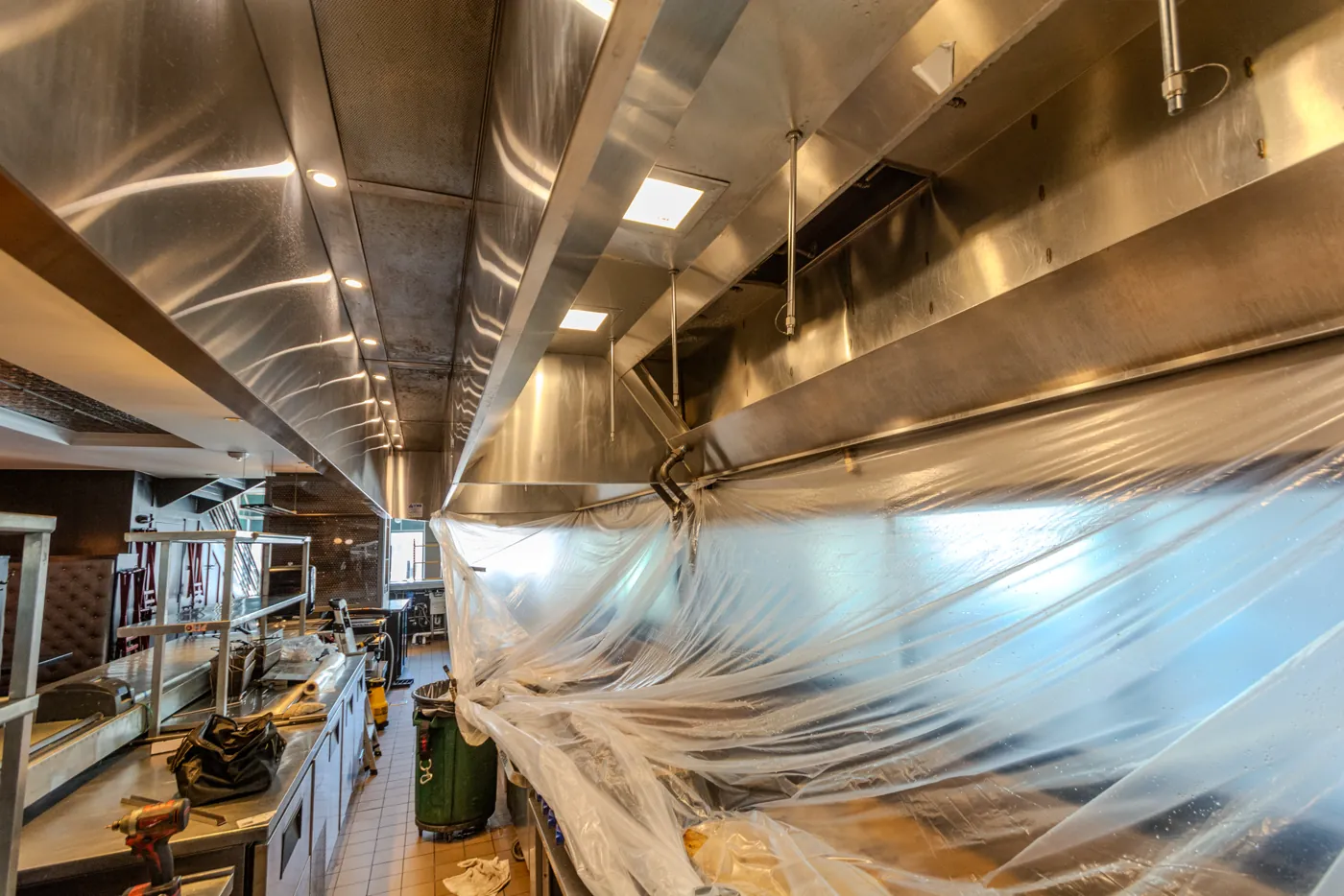A restaurant cook line sheeted in plastic from end to end, with the hood filters removed and the canopy exposed above it.