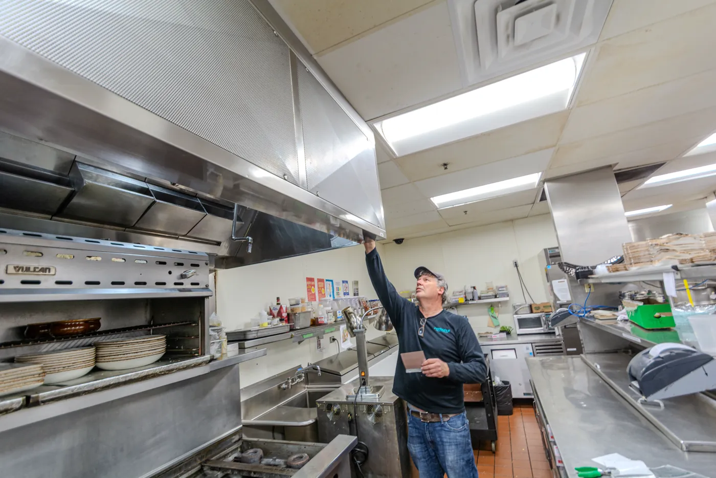 A technician in a Pinnacle shirt reaching up to a kitchen hood, checking the canopy with notes in hand.
