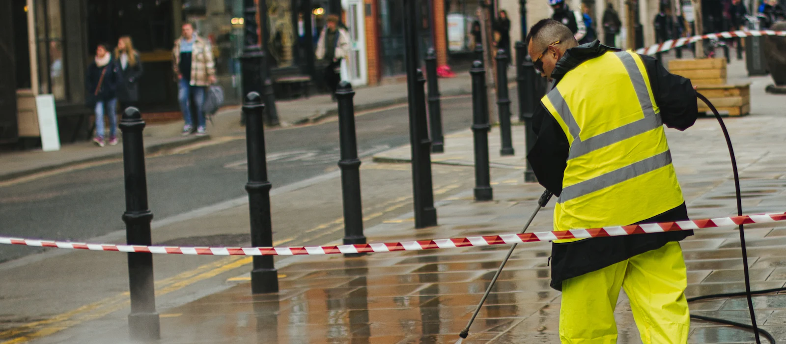 Worker in high-visibility gear pressure washing a wet city sidewalk behind a taped-off barrier.