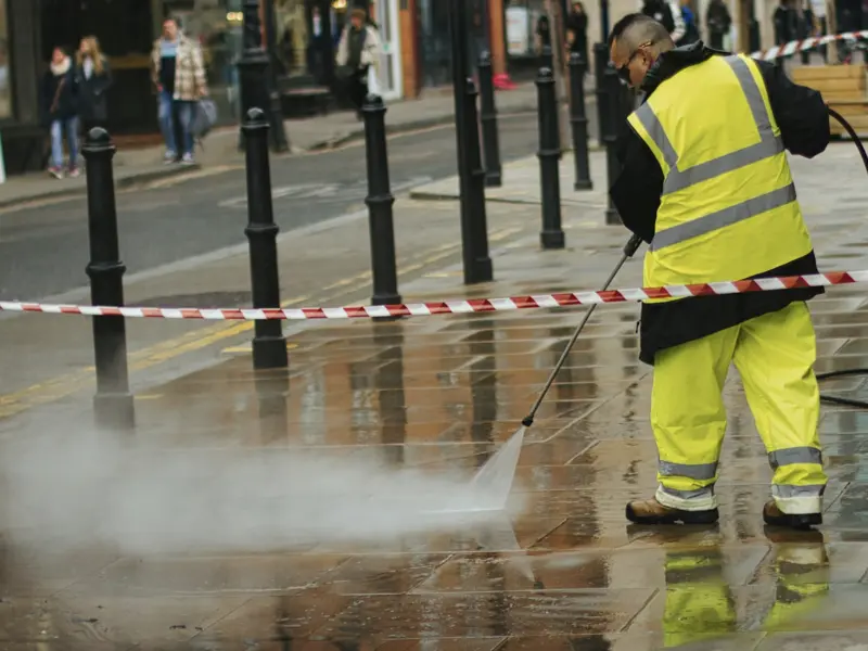 Worker in high-visibility gear pressure washing a wet sidewalk behind a taped-off barrier.
