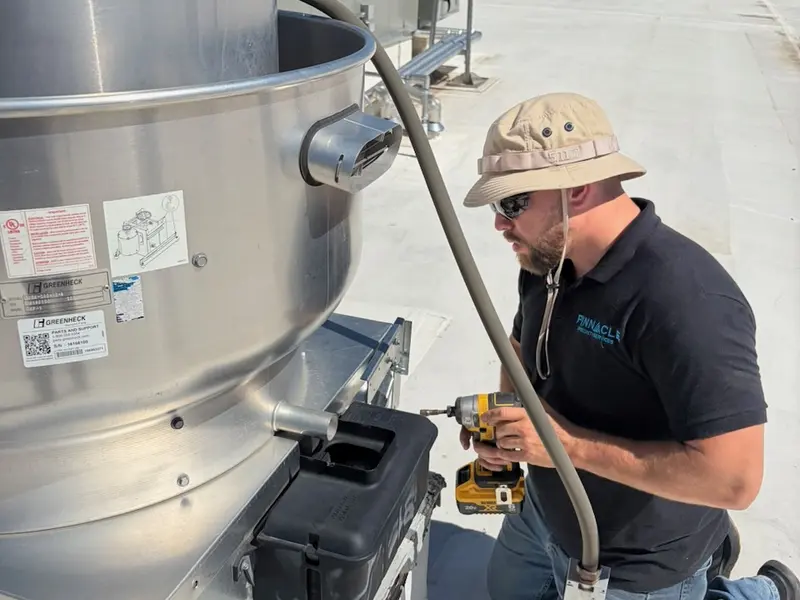 Technician in a sun hat kneeling at a rooftop exhaust fan housing, drill in hand.