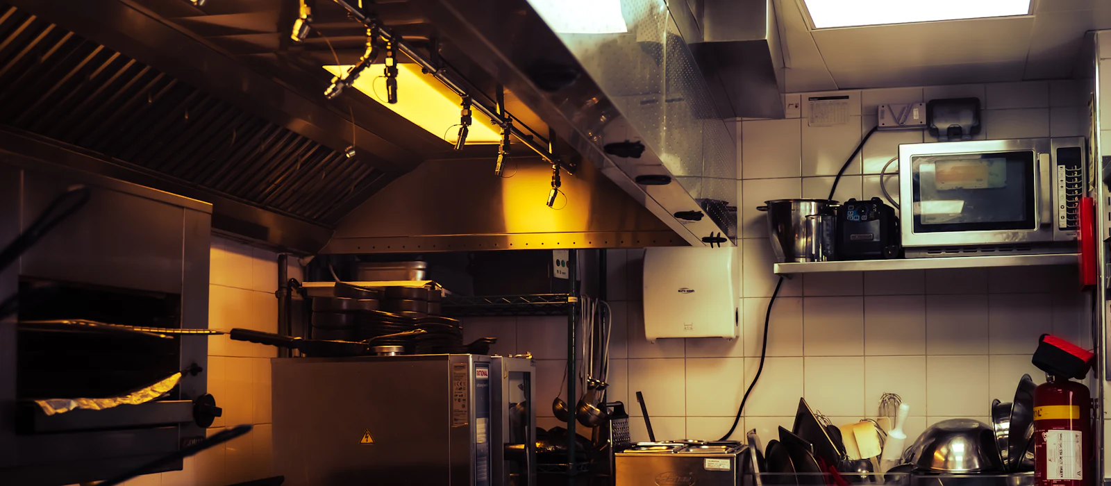 Stainless steel hood canopy over a commercial cooking line, lit by warm overhead lights.